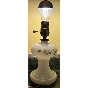 Könnte beinhalten: Weiße Keramik-Tischlampe mit einem klaren Glasschirm und einer eingeschalteten Glühbirne. Die Lampe hat ein florales Design auf dem Sockel.