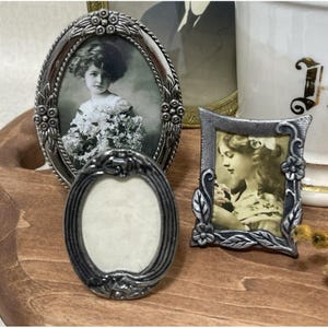 May include: An assortment of antique silver-toned picture frames in various shapes and sizes. One oval frame holds a vintage black and white photograph of a young girl. Another frame displays a portrait of a woman. The frames are arranged on a wooden surface.