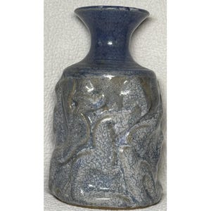 Puede incluir: Un pequeño jarrón de cerámica hecho a mano con un diseño orgánico texturizado. El jarrón presenta un cuello azul oscuro y un cuerpo con un esmalte moteado gris-azul. La superficie tiene una calidad rugosa y táctil.