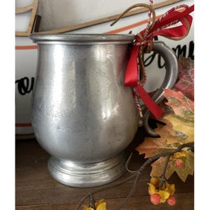 May include: A silver-coloured metal mug with a handle, tied with a red ribbon and twine. The mug has a bulbous body and a pedestal base. Autumn leaves and berries are in the background.