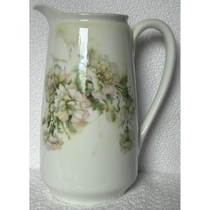 May include: White porcelain pitcher with a floral design. The pitcher has a wide mouth and a handle. The flowers are pink and white with green leaves.