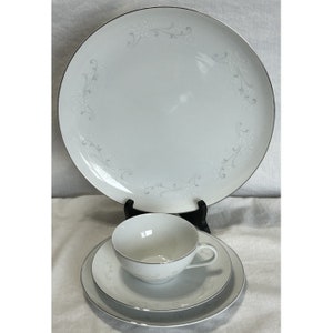May include: A set of white porcelain dinnerware featuring a delicate grey floral pattern and silver trim. The set includes a large dinner plate, a smaller salad plate, and a teacup and saucer.