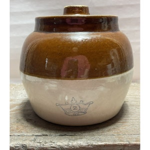 Puede incluir: Un tarro de cerámica esmaltado en marrón y blanco con tapa. El tarro tiene una corona con el número "2" en el interior en la parte delantera.