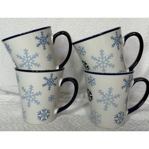 May include: Four white ceramic mugs with blue snowflake designs. Each mug has a black handle and a blue rim. The mugs are stacked and arranged on a white surface.