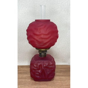 May include: A vintage ruby red oil lamp with a frosted glass globe and base. The lamp has a clear glass chimney and a brass-colored metal burner. The base is square-shaped, and the globe is round with a textured surface.