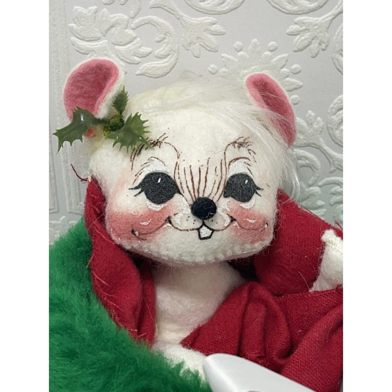 May include: A white plush mouse wearing a red and green coat with a holly sprig on its head. The mouse has black eyes and a painted smile.