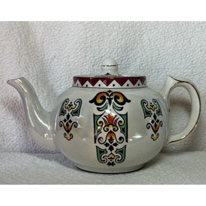 May include: White teapot with a geometric design in red, green, yellow, and orange. The teapot has a gold trim around the spout and handle.