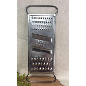 May include: A vintage metal grater with a rectangular design and a handle. The grater features multiple rows of small holes and two angled grating surfaces. The word "BROMCO" and "MADE IN U.S.A." are stamped on the front.