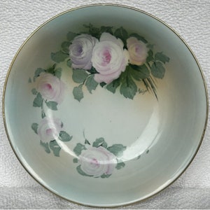 May include: A light blue ceramic bowl with a gold rim, featuring a hand-painted design of pink and purple roses with green leaves. The bowl has a signature on the bottom.