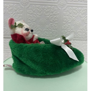 May include: A white mouse plush toy wearing a red scarf and a green holly wreath sits inside a green fuzzy slipper with a white bow and red berries.