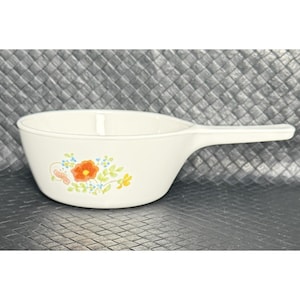 May include: Off-white ceramic serving bowl with a handle. The bowl features a floral design with orange, yellow, and blue flowers. The handle extends from the side of the bowl, providing a comfortable grip. This bowl is ideal for serving soups and stews.