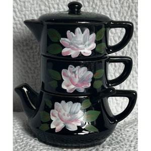 Puede incluir: Una tetera de cerámica negra con tapa y dos tazas apiladas encima. La tetera y las tazas están decoradas con flores rosas y blancas pintadas a mano y hojas verdes.