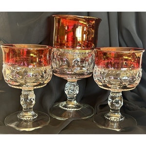 Puede incluir: Conjunto de tres copas de vidrio rojo rubí con base de vidrio transparente y un intrincado diseño de vidrio cortado. Las copas están sobre un fondo de tela marrón oscuro.