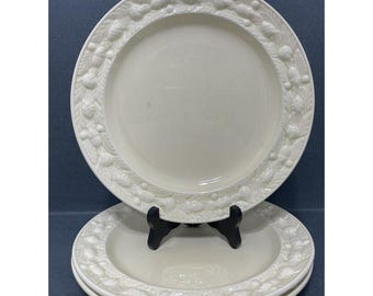 May include: Off-white ceramic plates with a raised acorn and leaf border. The plates are stacked on a black plate stand. The plates have a simple, elegant design, suitable for everyday use or special occasions.