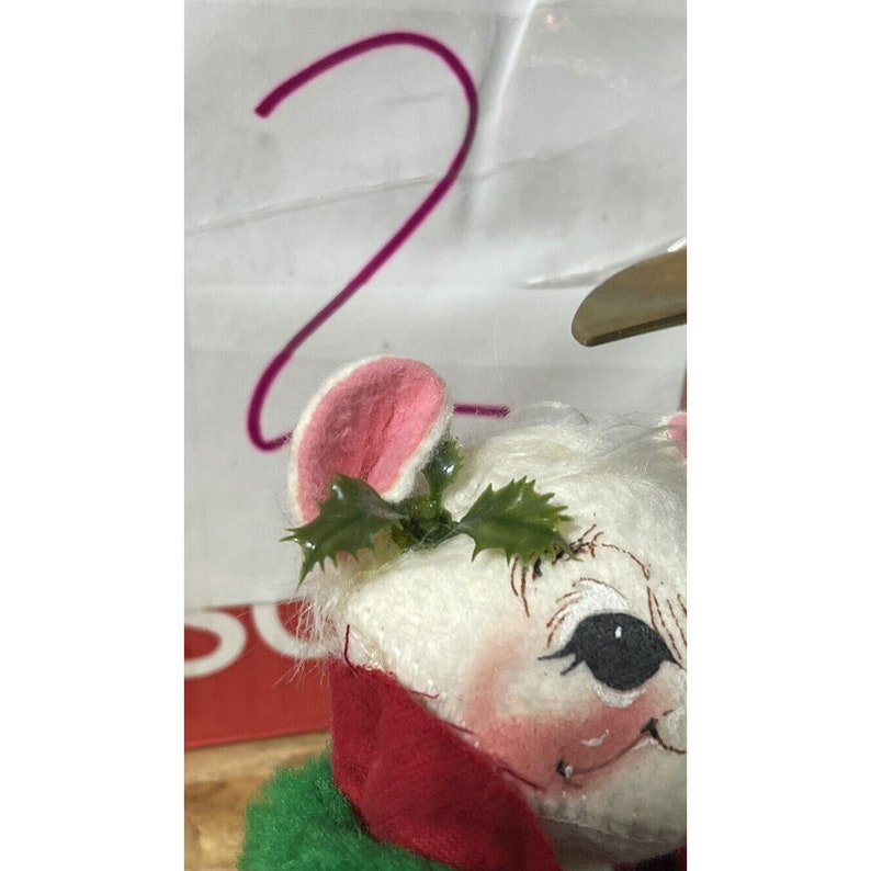 May include: A white plush mouse wearing a red and green outfit with a holly sprig on its head. The mouse has a pink nose and black eyes.