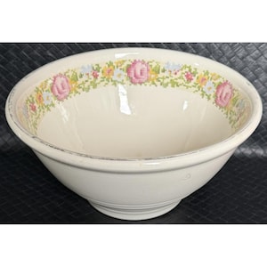 Pode incluir: Tigela de cerâmica creme com um desenho floral em ponto cruz ao redor da borda. A tigela tem uma forma larga e arredondada e uma borda ligeiramente desgastada. O padrão floral inclui rosas cor-de-rosa, flores amarelas e azuis.