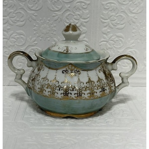 Puede incluir: Un azucarero de porcelana blanca con tapa, con un delicado diseño floral verde y dorado. El tazón tiene dos asas y un borde festoneado.