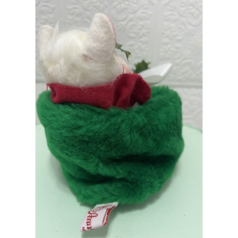 May include: A white plush Christmas gnome wearing a red scarf and sitting in a green plush stocking. The gnome is holding a small white object in its hand. The stocking has a red and white "Anna's" label.