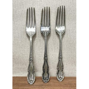 May include: Three antique silver forks with ornate floral detailing on the handles. The forks are arranged vertically against a neutral background, showcasing their tines and decorative elements. The silverware is likely vintage.