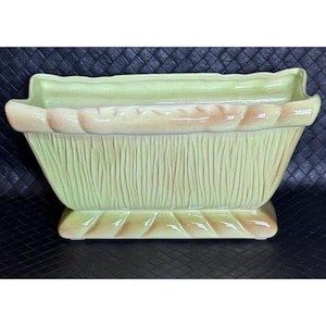 May include: A light green ceramic planter with a textured, woven design. The planter has a rectangular shape with rounded edges and a slightly raised base.