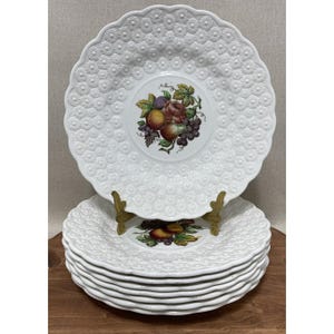 May include: A stack of white ceramic plates with a scalloped edge and embossed floral pattern. Each plate features a central fruit and floral design. The plates are displayed on a wooden surface.