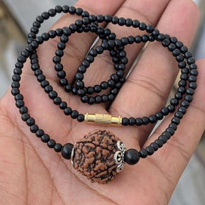 Puede incluir: Collar de cuentas negras con un colgante central marrón texturizado. El collar presenta un cierre dorado y una cuenta decorativa plateada. Las cuentas son pequeñas y redondas, creando un efecto en capas.