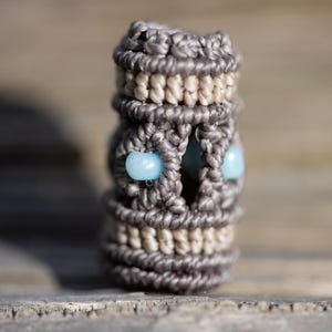 Könnte beinhalten: Eine graue und beige Makramee-Perle mit zwei hellblauen Perlen. Die Perle ist mit einer Knotentechnik hergestellt.