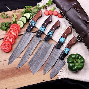 May include: A set of five Damascus steel kitchen knives with wooden handles and turquoise accents. The blades are arranged on a wooden surface with sliced tomatoes, cucumbers, and a green bell pepper. A brown leather knife case is visible.