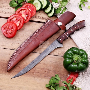 May include: A Damascus steel knife with a wooden handle and a brown leather sheath. The knife is next to sliced tomatoes, cucumbers, and a green bell pepper. The knife has a decorative pattern on the blade.