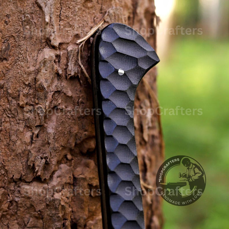 K&ouml;nnte beinhalten: Nahaufnahme eines schwarzen, geschnitzten Holzgriffs mit einem strukturierten, wabenartigen Muster. Ein kleiner silberner Niet ist oben zu sehen. Der Griff ist vor einem rauen, braunen Baumstamm platziert. Das ShopCrafters-Logo ist sichtbar.