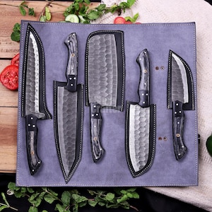 May include: A set of five kitchen knives with hammered metal blades and dark wood handles, each stored in a black leather sheath. The knives are arranged on a light purple leather roll-up mat, featuring a variety of blade shapes and sizes.