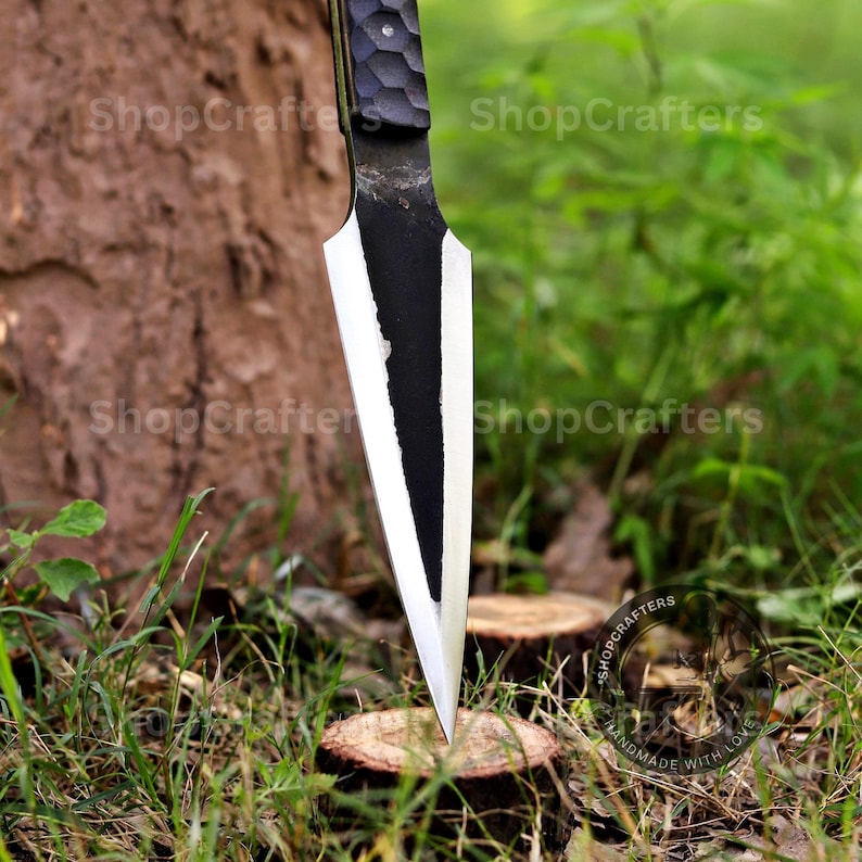 K&ouml;nnte beinhalten: Ein handgefertigtes schwarzes und silbernes Messer mit strukturiertem Griff steckt in einem Baumstumpf. Die Klinge ist scharf und spitz, mit einer kontrastierenden silbernen Kante. Das Messer ist von gr&uuml;nem Gras und Bl&auml;ttern umgeben.