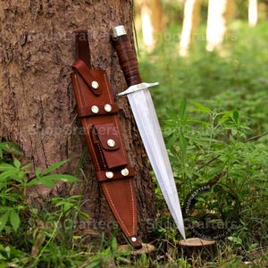 May include: A dagger with a polished steel blade and a brown leather-wrapped handle rests against a tree. The dagger is accompanied by a brown leather sheath with decorative metal studs. The scene is set outdoors with green foliage.