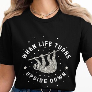 Puede incluir: Camiseta negra con un gráfico blanco de un perezoso colgado boca abajo con el texto "When life turns upside down".