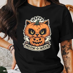 Può includere: Maglietta nera con una grafica di un gatto che indossa una zucca sulla testa. Il testo "Happy Meow-O-Ween" è stampato sotto il gatto.