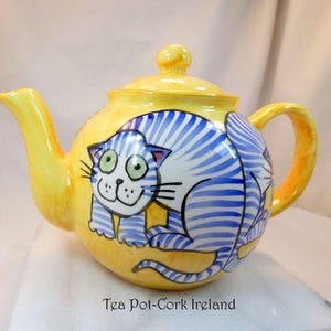 May include: A yellow ceramic teapot with a whimsical cat design. The cat is white with blue stripes and green eyes. The teapot has a curved spout, a rounded handle, and a lid with a small knob. The text "Tea Pot-Cork Ireland" is at the bottom.