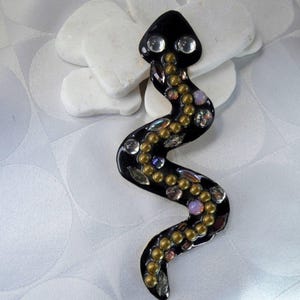 May include: A black snake-shaped decorative item with a glossy finish. The snake is adorned with clear, gold, and iridescent embellishments. The snake's head features two clear, round accents, and the body is curved. The item is placed on a white surface.