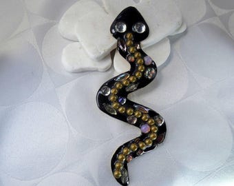 May include: A black snake-shaped decorative item with a glossy finish. The snake is adorned with clear, gold, and iridescent embellishments. The snake's head features two clear, round accents, and the body is curved. The item is placed on a white surface.