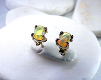 May include: A pair of stud earrings featuring rectangular, iridescent opal gemstones. The opals display a mix of yellow, orange, and green hues, set in silver-toned metal with a claw-like design. The earrings are displayed on a white, textured surface.