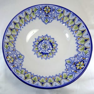 May include: A white ceramic serving bowl with a blue rim and intricate floral patterns in blue, green, and yellow. The design features a central floral medallion and a repeating border pattern. The bowl has a glossy finish.