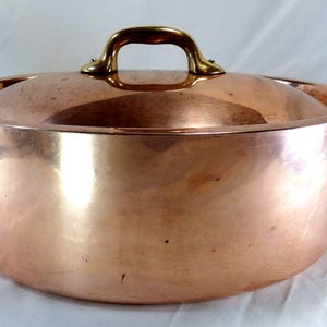 May include: A copper cooking pot with a lid and brass handles. The pot has a rounded shape and a shiny, reflective surface. The lid has a brass handle. The pot is designed for cooking and food preparation.