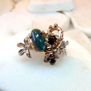 May include: A silver-toned ring featuring an oval, dark green gemstone and smaller, round, dark red gemstones. The ring has a floral design with intricate details and is set against a white background.