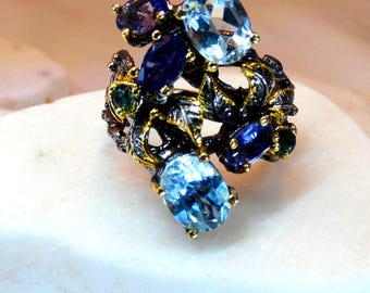May include: A statement ring featuring a cluster of blue and purple gemstones. The ring has a black and gold metal setting with a floral design. The gemstones include oval and pear-shaped stones in various shades of blue and purple.