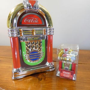 May include: A ceramic Coca-Cola jukebox cookie jar with red, green, and gold accents, featuring the text Drink Coca-Cola and Rock n Roll. Beside it is a smaller, boxed Coca-Cola jukebox salt and pepper set with the text Coke A Song on the packaging.
