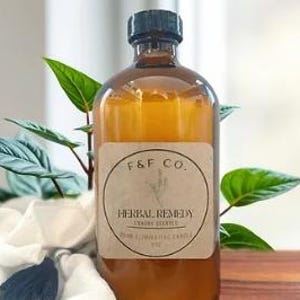 Pode incluir: Uma garrafa de vidro âmbar com tampa de rosca preta e um rótulo retangular bege. O rótulo exibe o texto F&F CO. no topo, seguido por uma ilustração botânica e as palavras HERBAL REMEDY, ODOR ELIMINATING CANDLE, 8 OZ.