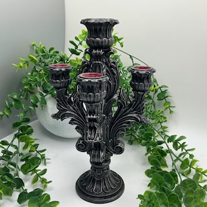 May include: A black candelabra with a distressed silver finish featuring four candle holders arranged in a tiered structure. The base and arms are decorated with ornate, carved leaf-like patterns and scrollwork details.