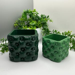 May include: Two square planters with a tufted button design. The larger planter is dark forest green, and the smaller planter is a lighter shade of green. Both containers feature a textured, padded appearance with recessed buttons on all sides.