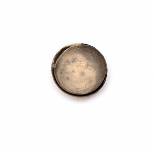 May include: A circular button with a light beige, textured face and a dark, metallic rim. The button has a mottled appearance, suggesting age or wear. The button is set against a plain white background. The button is approximately 2.5 cm in diameter.