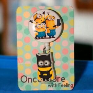 May include: A badge reel featuring Minions characters and a Batman-themed Minion charm. The reel has a round image of three Minions on a white background. The charm is a Minion dressed as Batman. The background is a card with colorful polka dots.