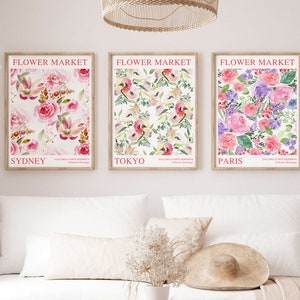 May include: Three framed floral art prints with the text "Flower Market" above the city names Sydney, Tokyo, and Paris. The prints feature watercolor floral patterns in pink, purple, and green hues. The prints are displayed above a white sofa.